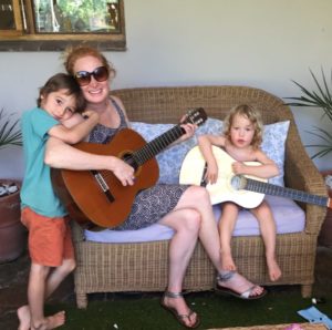 Astrid pictured seated on a wicker loveseat with two young children, and holding guitars.